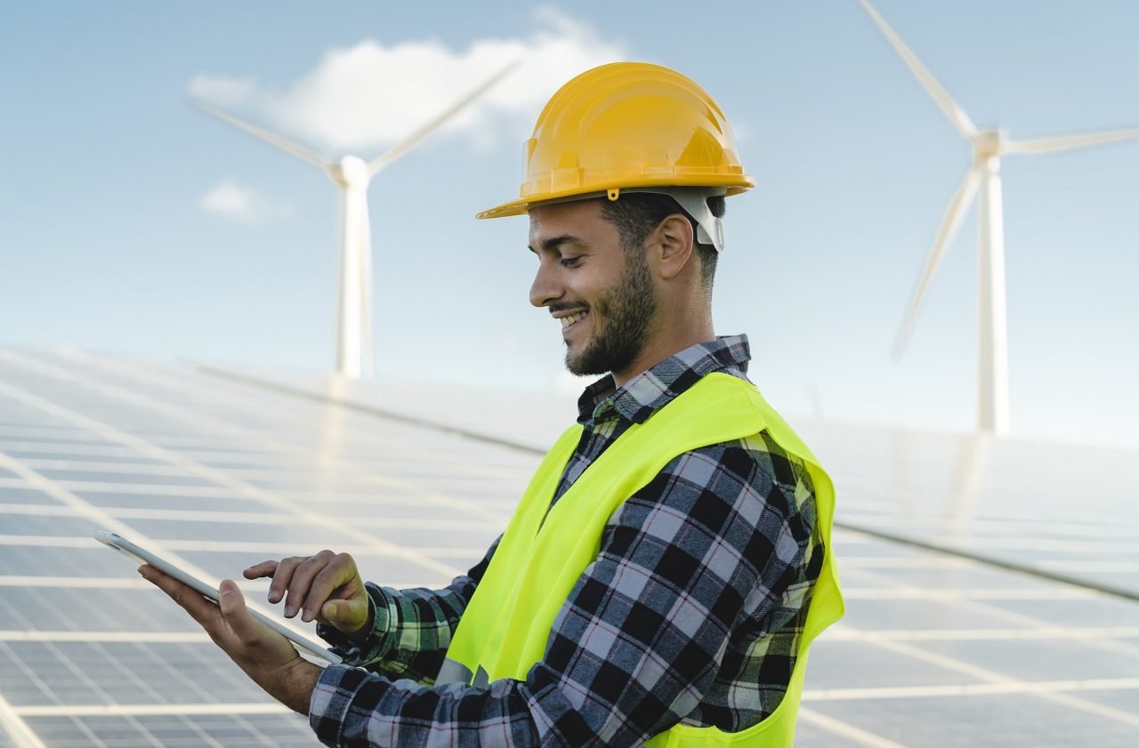 An engineer in a safety vest and helmet reviewing blueprints near a solar panel installation site managed by AP Sun Energy, ensuring project efficiency and quality.